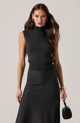 A woman is wearing a sleeveless, dark gray turtleneck top and a matching high-waisted, long skirt. She accessorizes with gold hoop earrings, a large gold bracelet, and a black handbag.